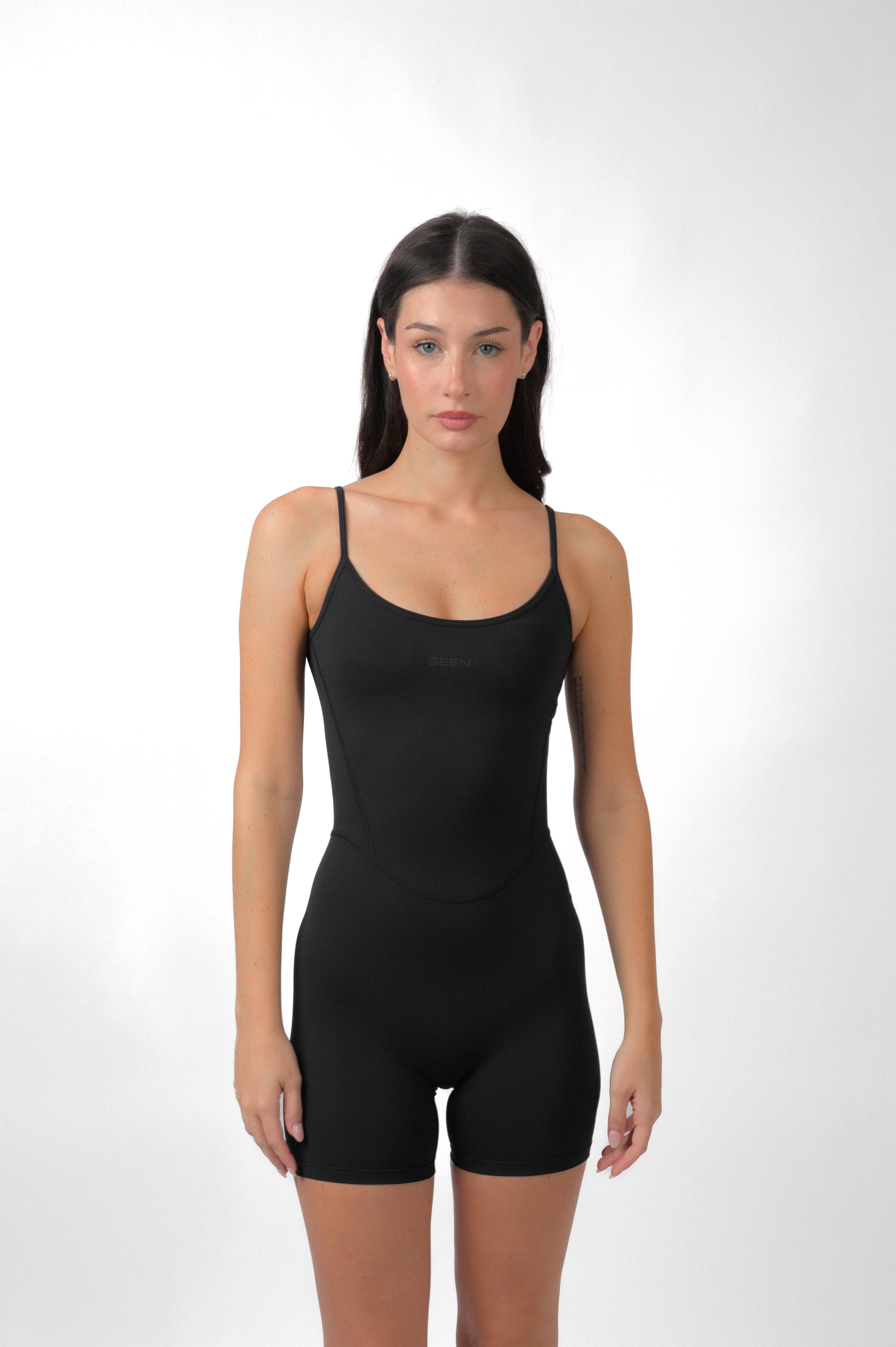 A female model wearing a black strappy romper with a low scoop neckline and adjustable straps.